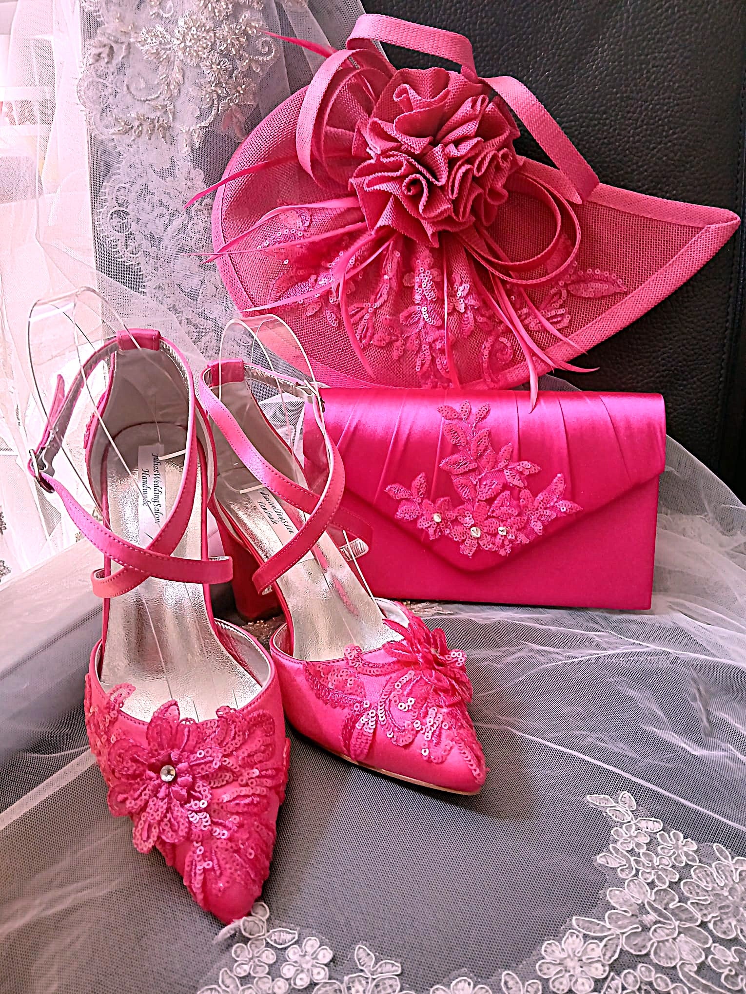 A coordinated pink bridal accessory set including a pair of pointed-toe high heels with floral lace and ribbon embellishments, a matching fascinator hat, a satin clutch, and a large matching fascinator flower arrangement, arranged on sheer tulle fabric.